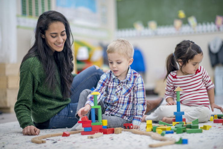 A preschool boy and girl are indoors with their teacher. They are sitting on the floor and playing with colorful blocks together.